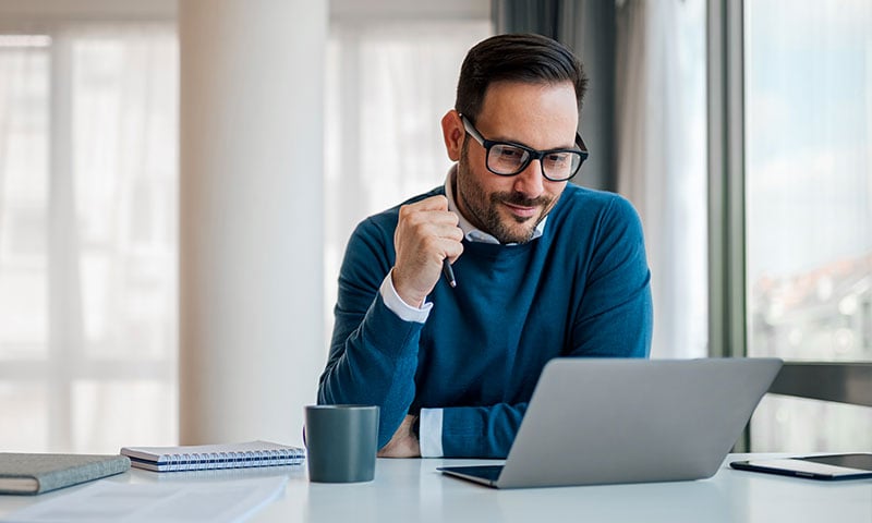 Professional man wearing glasses, sitting in front of a laptop with a mug and notebook next to him